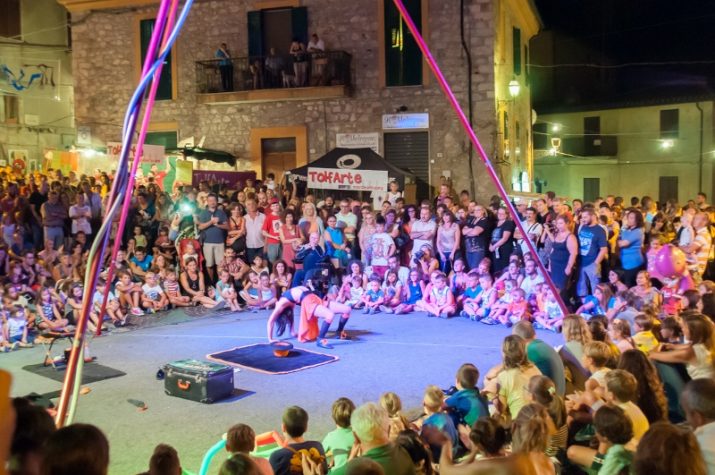 Spettacolo serale a TolfArte con la piazza gremita di spettatori, artisti sul palco e giochi di luce tra le vie del centro storico