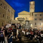 Veduta serale di Piazza Santa Maria in Trastevere a Roma con la basilica illuminata, la fontana e i tavolini all'aperto durante una serata estiva.